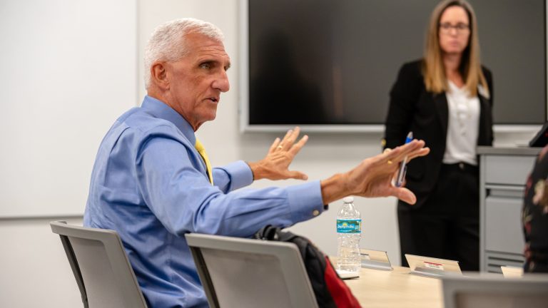 Mark Hertling gestures while speaking, seated at a classroom table, with a woman standing near a presentation screen in the background.