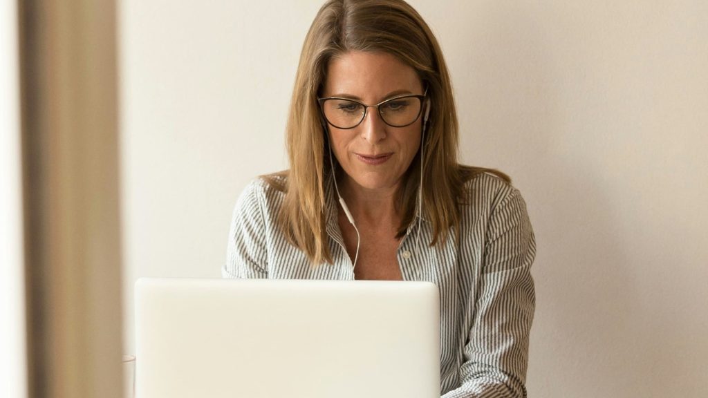 A focused woman wearing glasses and earbuds working on her laptop.